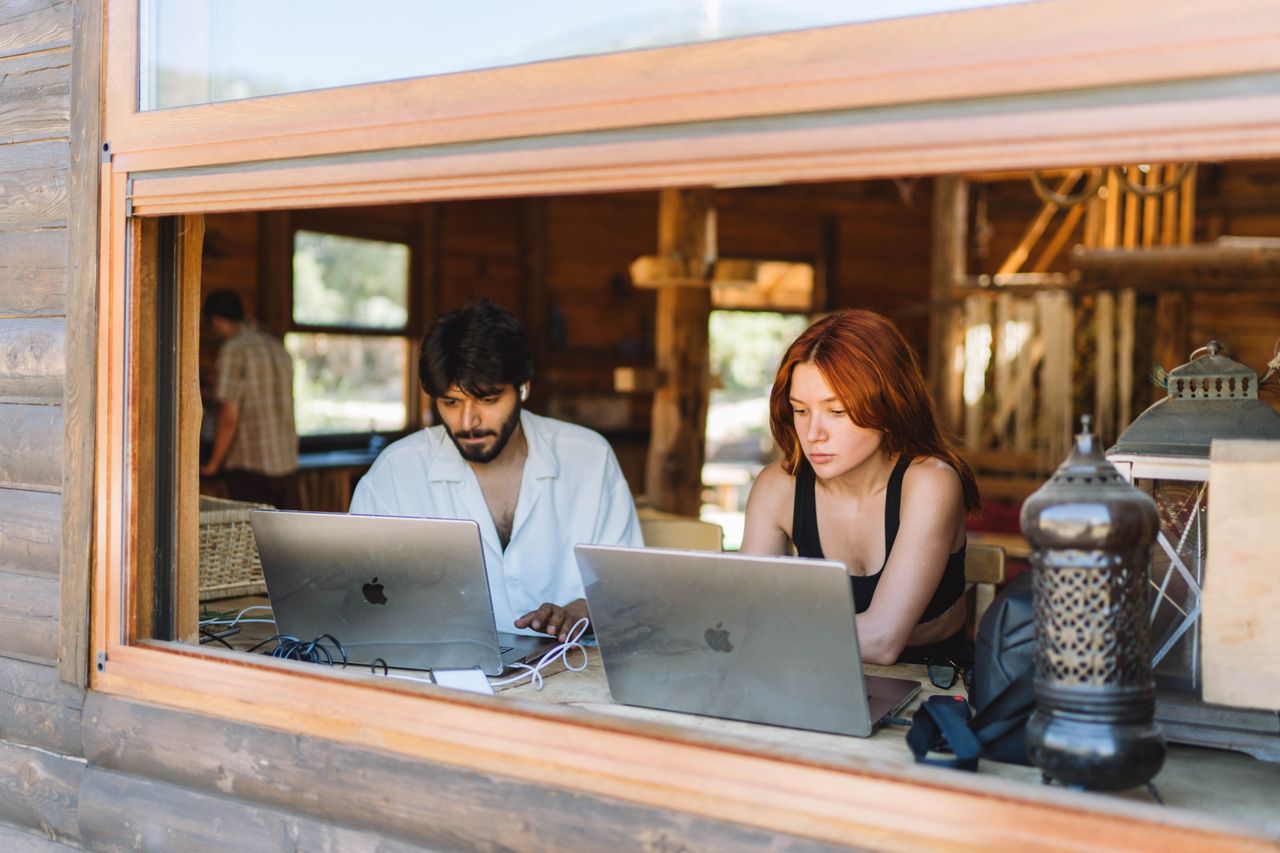 Man and woman collaborating on laptops by a large window in a coworking area